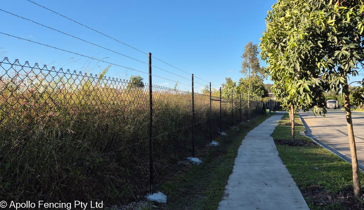 Black PVC-coated chain wire fencing with barbed wire installed along the industrial boundary of a grassy block in the Gold Coast. A footpath runs parallel to the fence, lined with trees and a clear blue sky above.